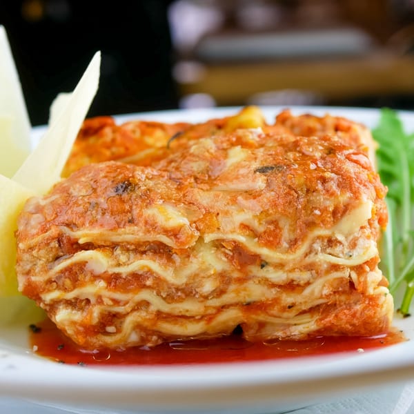 Close-up of a slice of lasagna with rich red sauce, cheese, and fresh greens on a white plate.
